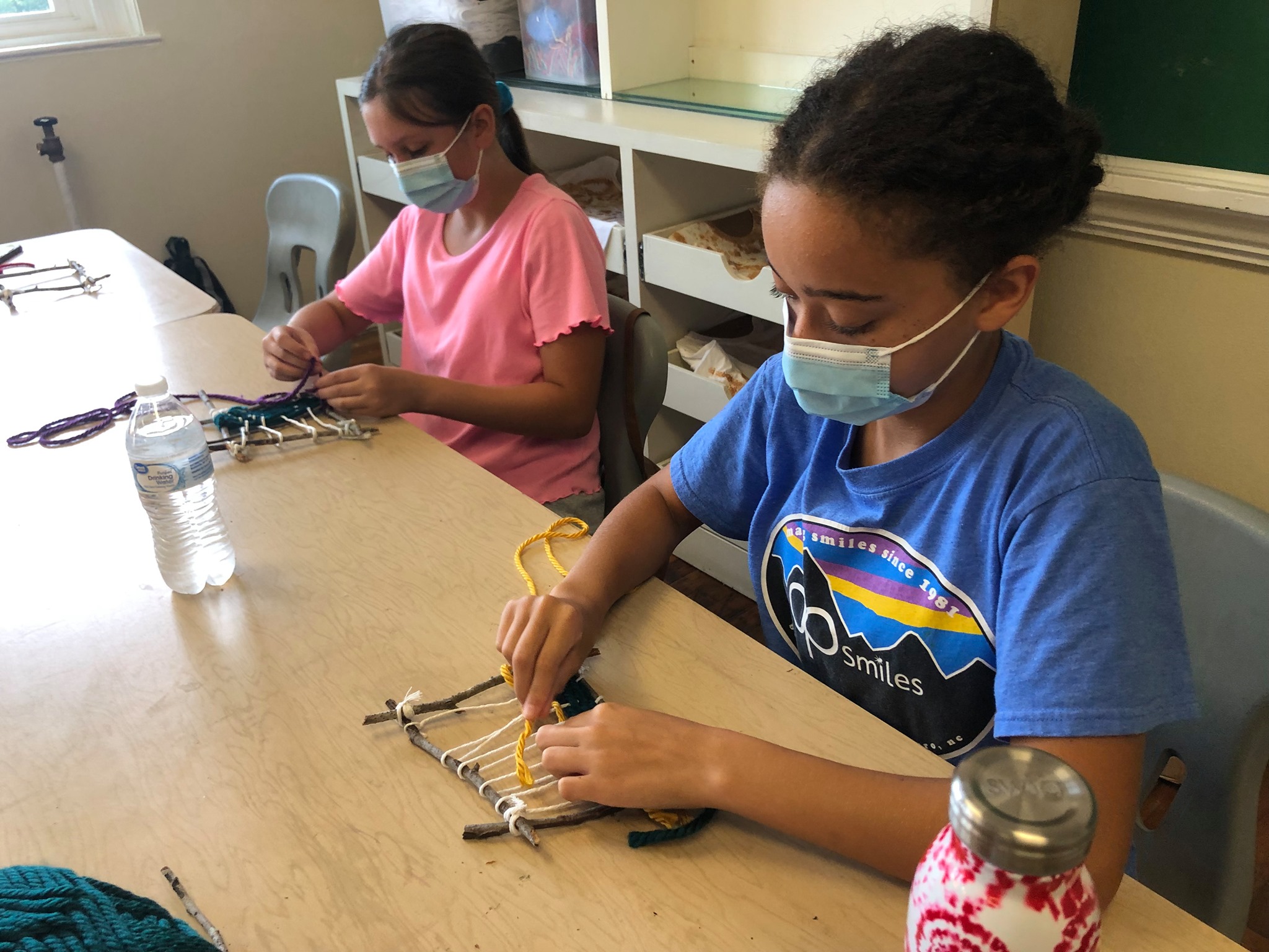 Using sticks and string to make a small loom, Zoe Woods (right) and Sierra Wood (left) weave thick yarn through their warp during Art Outside in July.